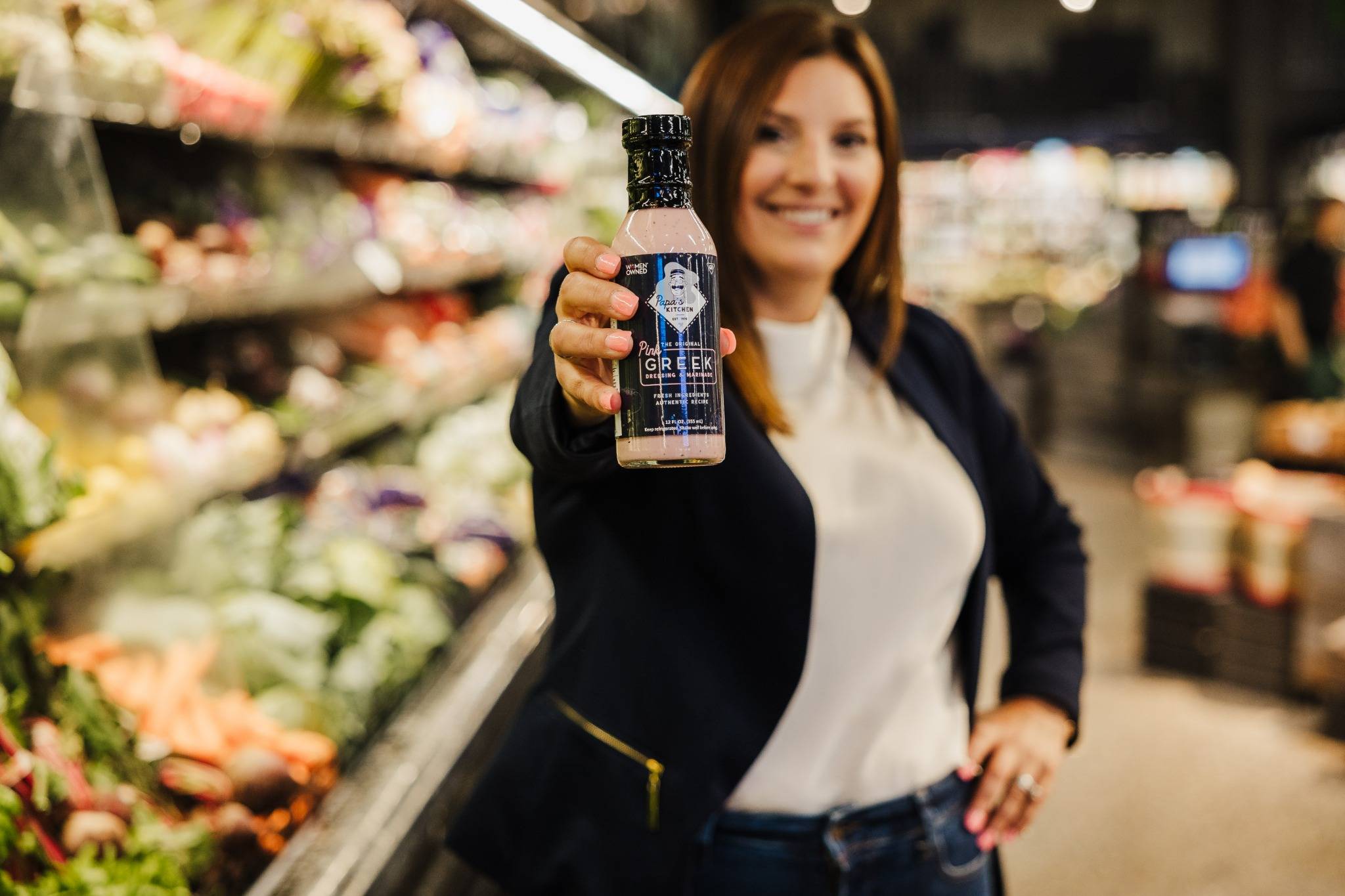 Christina Kalfaian, founder of Papa’s Kitchen, holding a bottle of Pink Greek™ dressing inside a grocery store produce aisle.
