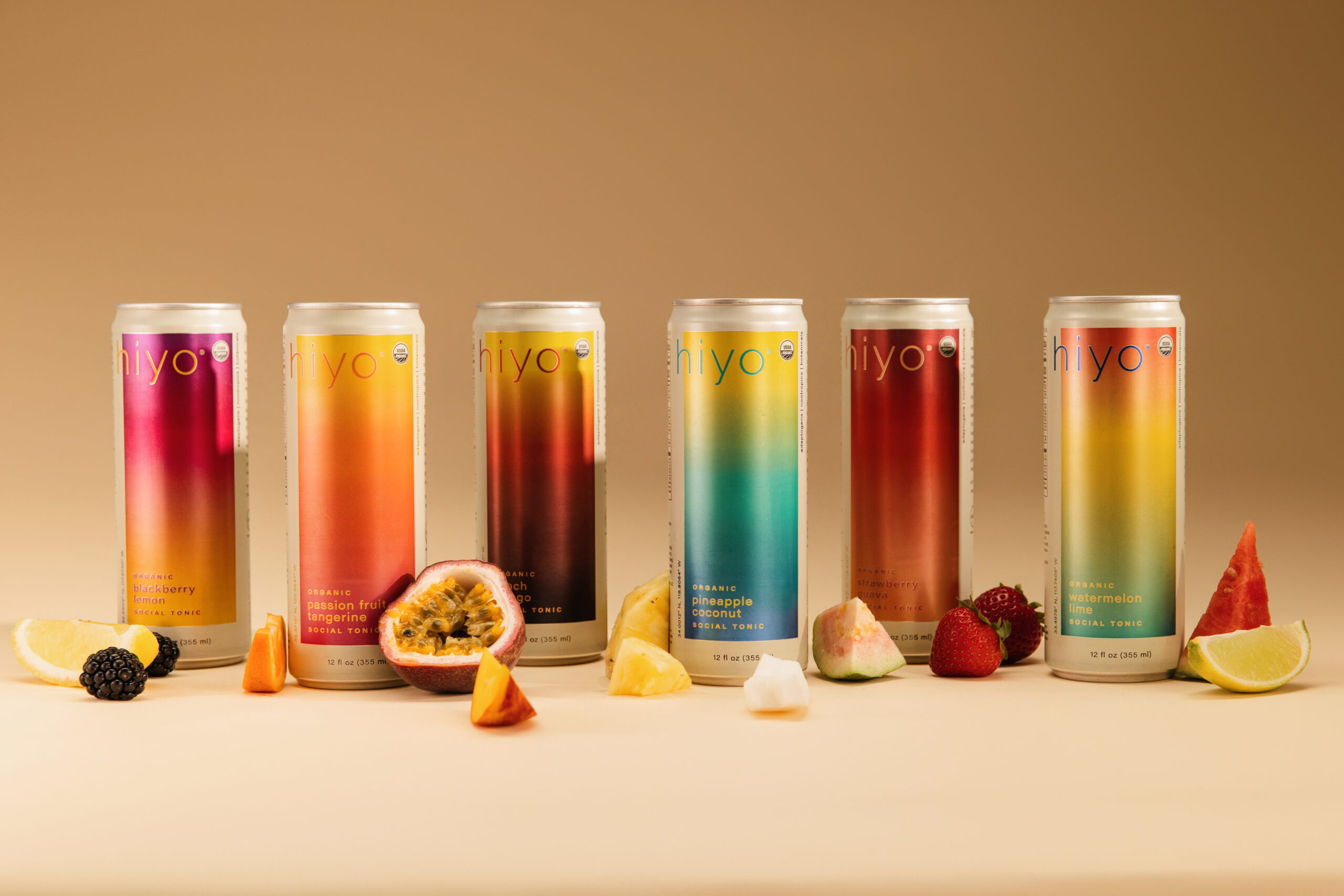 Six Hiyo social tonic cans arranged in a row with fresh fruit pieces in front, displayed on a neutral backdrop.
