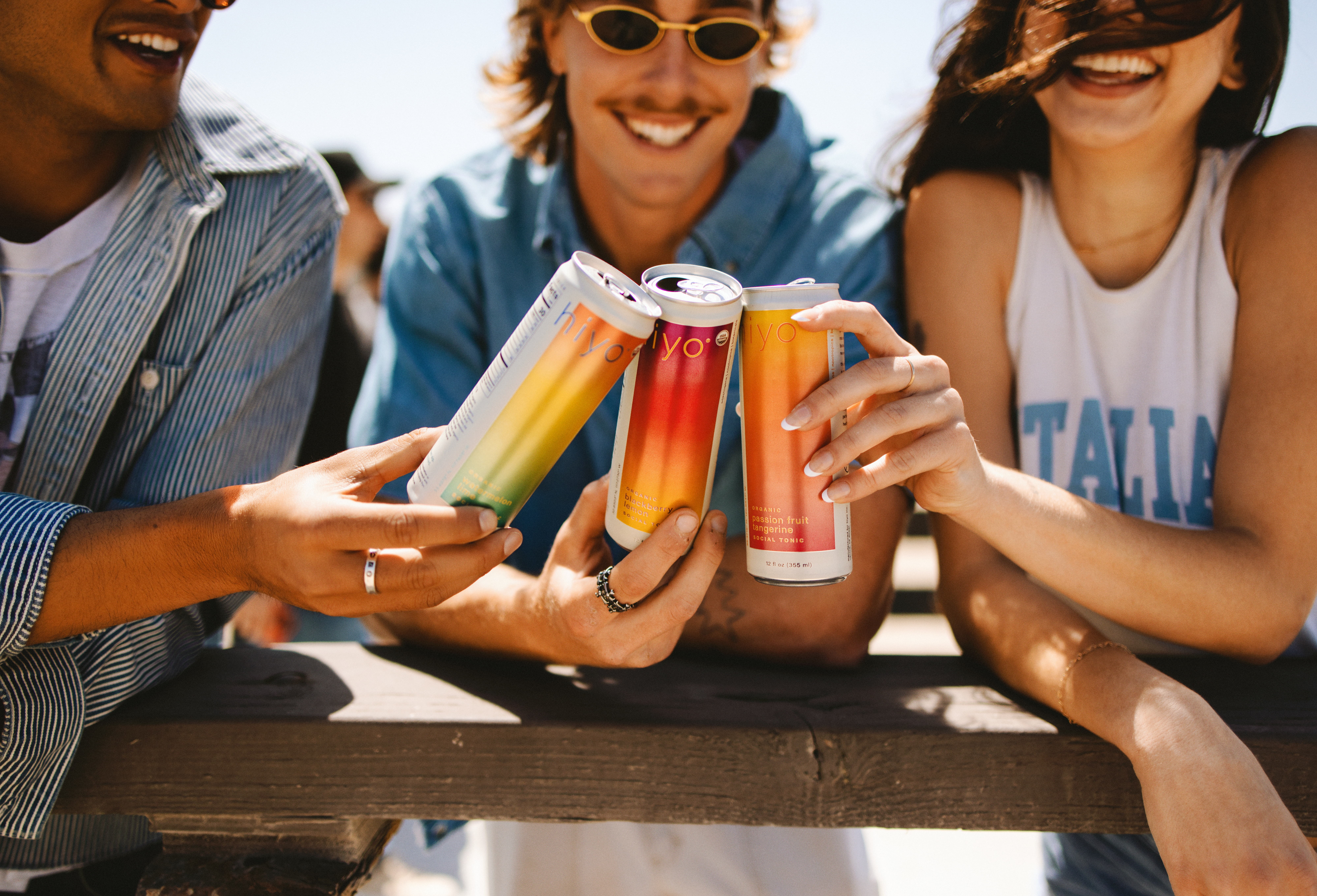 Three people outdoors clinking Hiyo cans together, smiling and enjoying a sunny moment.