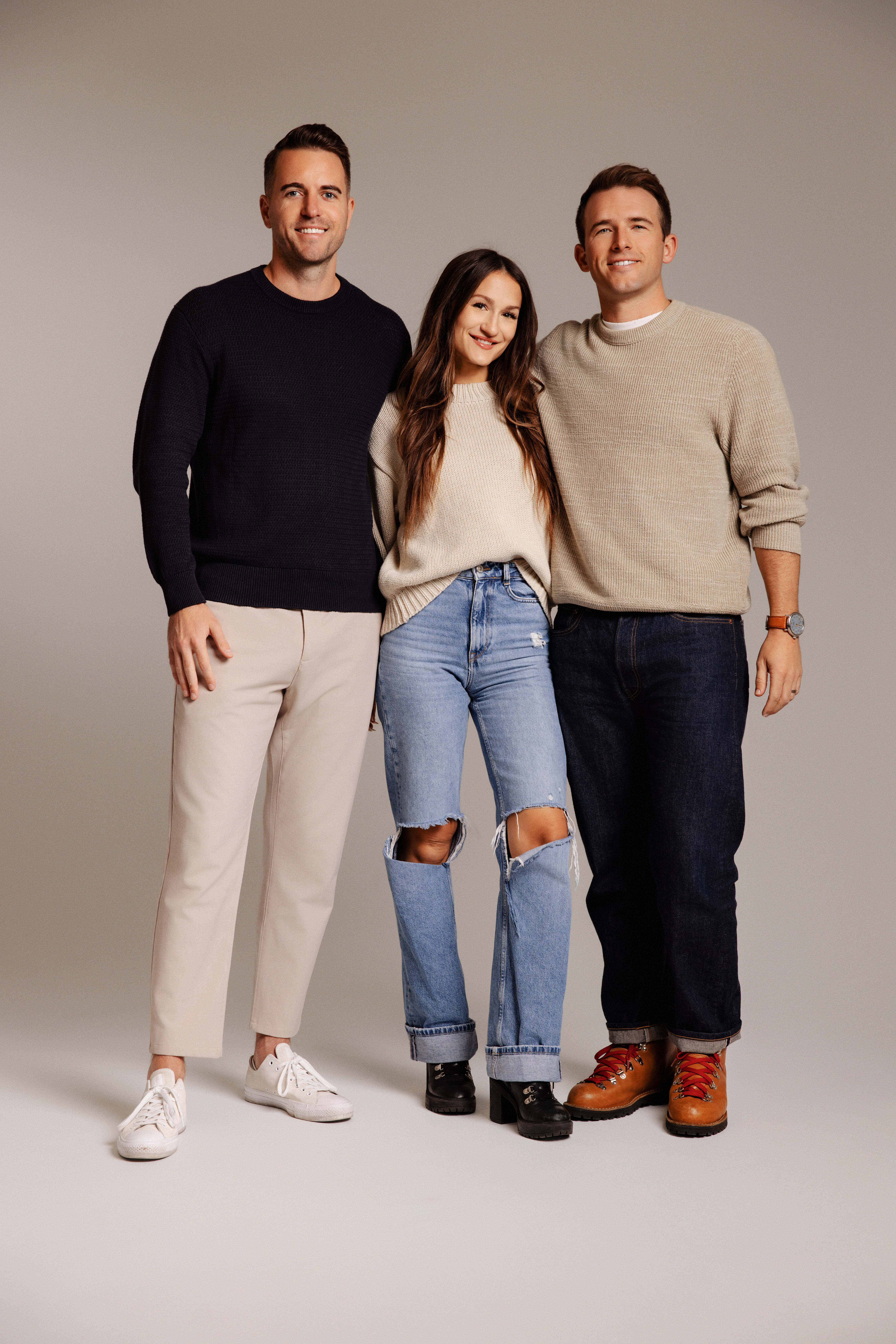 Three team members standing together in a studio, wearing casual sweaters and jeans, smiling at the camera.