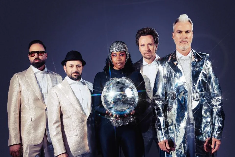 Fitz and The Tantrums band members stand shoulder‑to‑shoulder against a dark studio backdrop, wearing coordinated formal and metallic outfits. The person at center holds a mirrored disco ball that reflects light across the scene, creating a dramatic atmosphere.